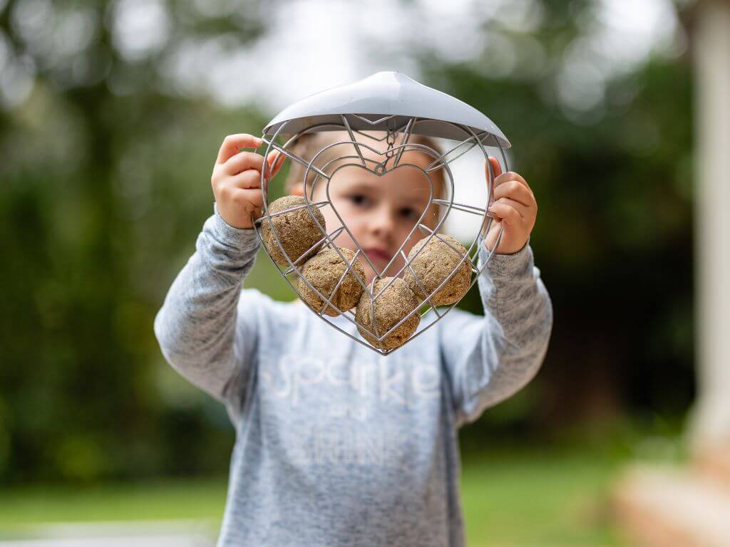 A child holding a heart-shaped suet feeder filled with suet balls, with greenery blurred in the background.