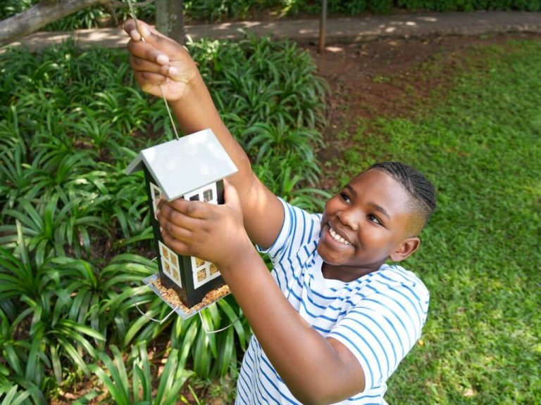 A smiling boy hangs a black and white Westerman’s house feeder filled with seed in a leafy garden.