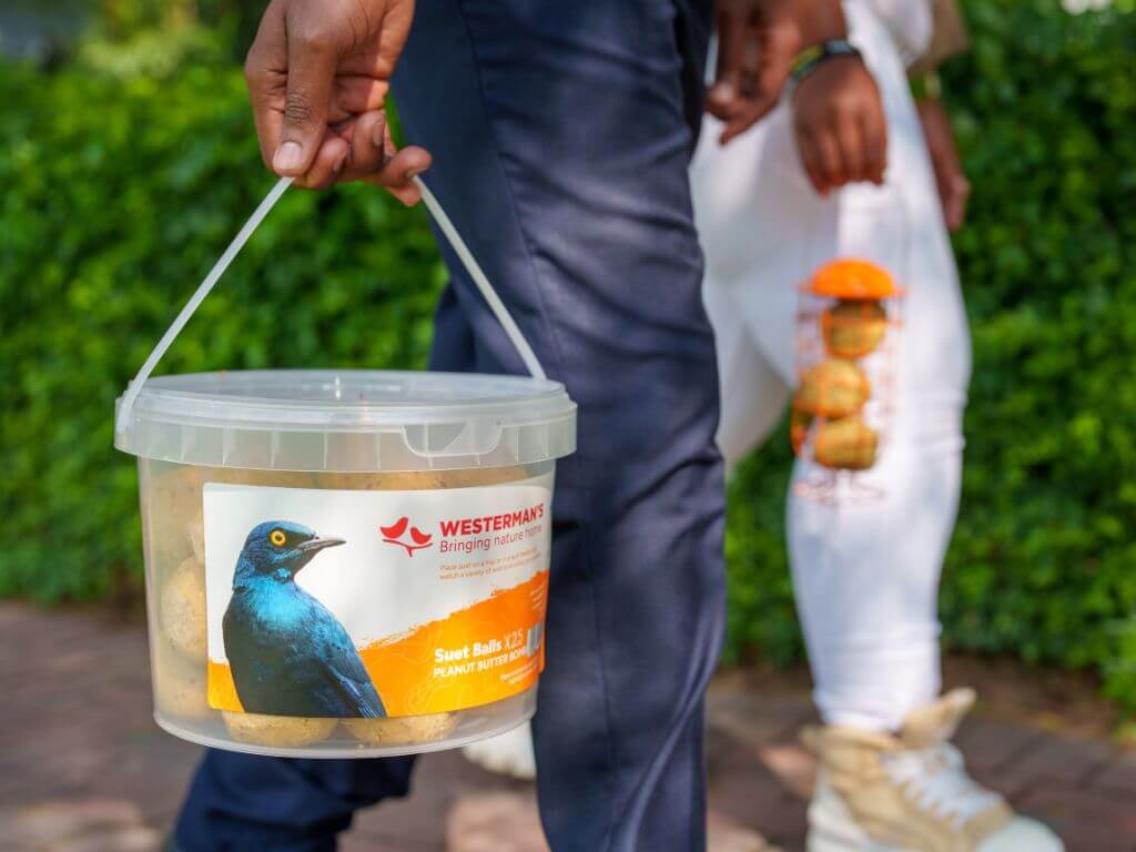 A person carries a tub of Westerman’s Peanut Butter suet balls while another holds a suet ball feeder in the background.