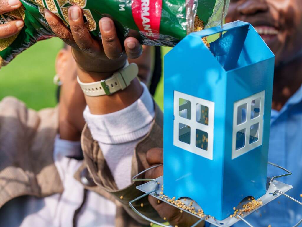 Two adults pour Westerman’s bird seed into a bright blue house feeder outdoors.