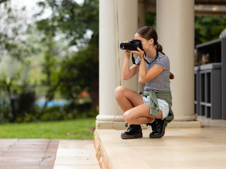 A young girl crouches with a DSLR camera, preparing to take bird photos from a shaded patio.