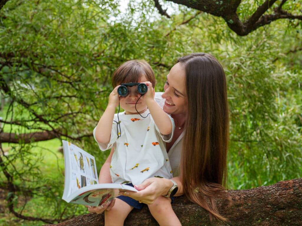 A smiling toddler looks through binoculars while sitting on a tree branch with their mother and a bird book.