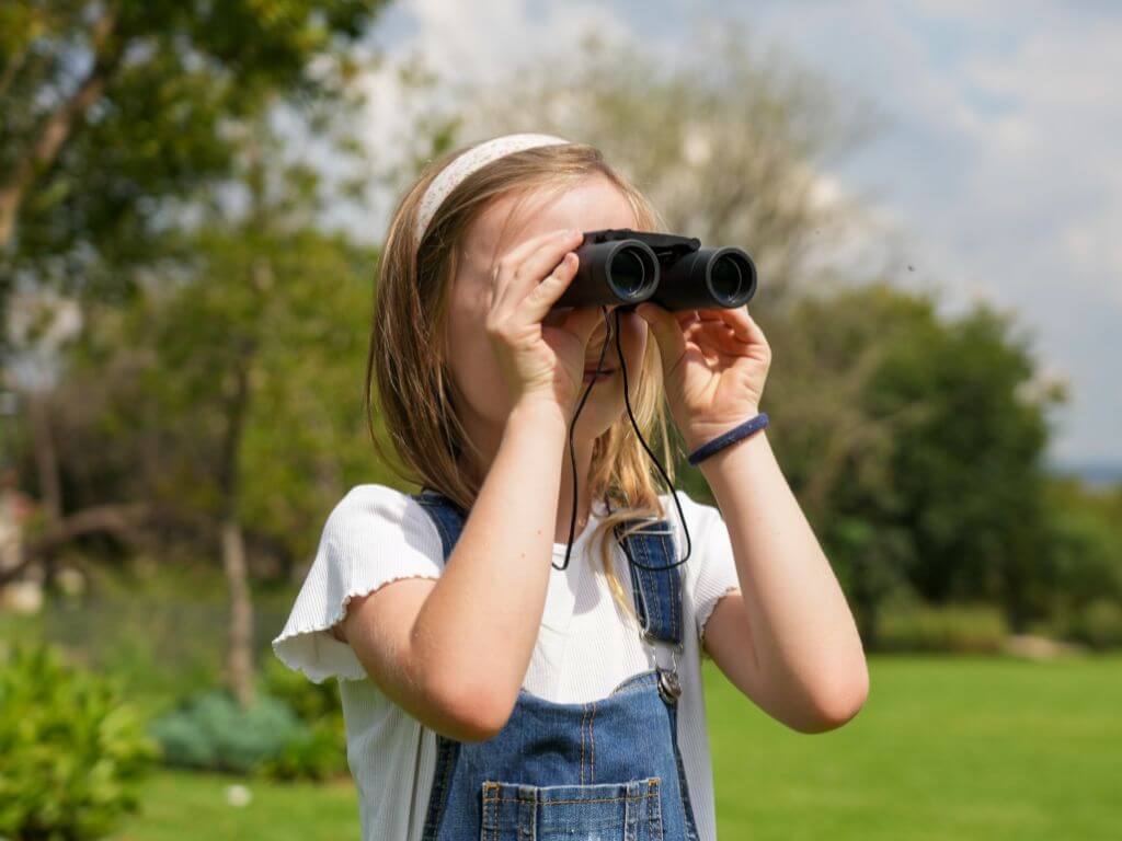 A young girl wearing a denim pinafore uses binoculars to spot birds in a sunny garden setting.