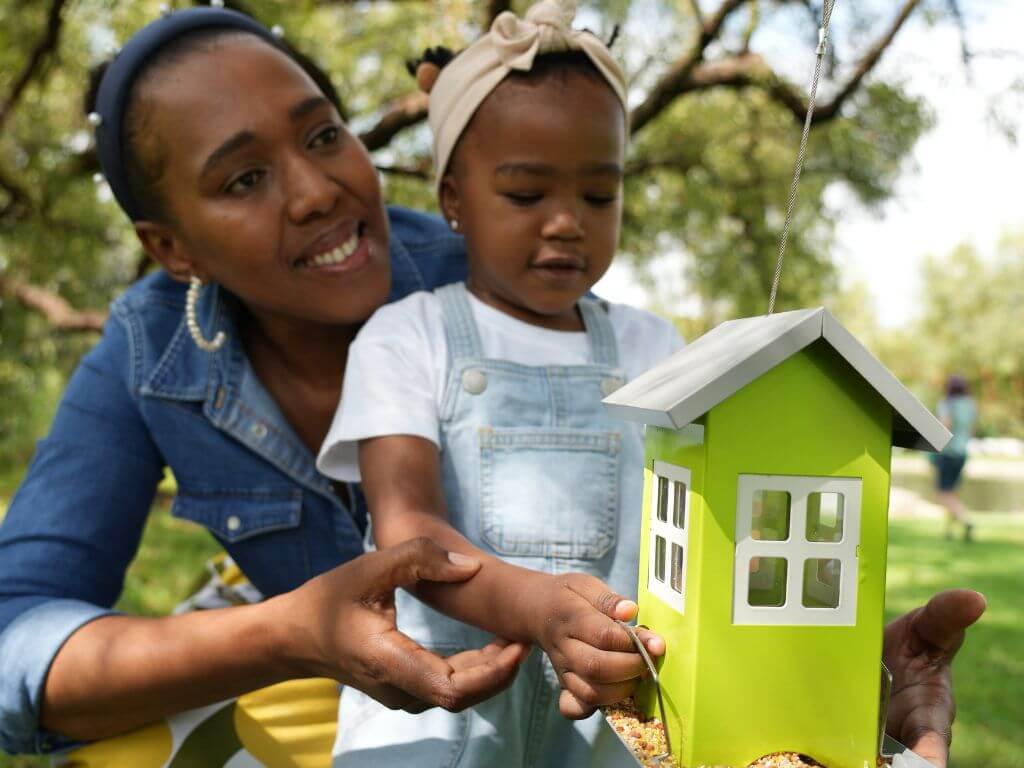 A mother helps her young daughter hang a green Westerman’s house feeder filled with bird seed.