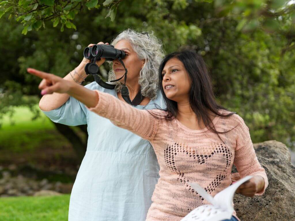 A woman points while another uses binoculars to spot birds, both surrounded by greenery and holding a bird guide.
